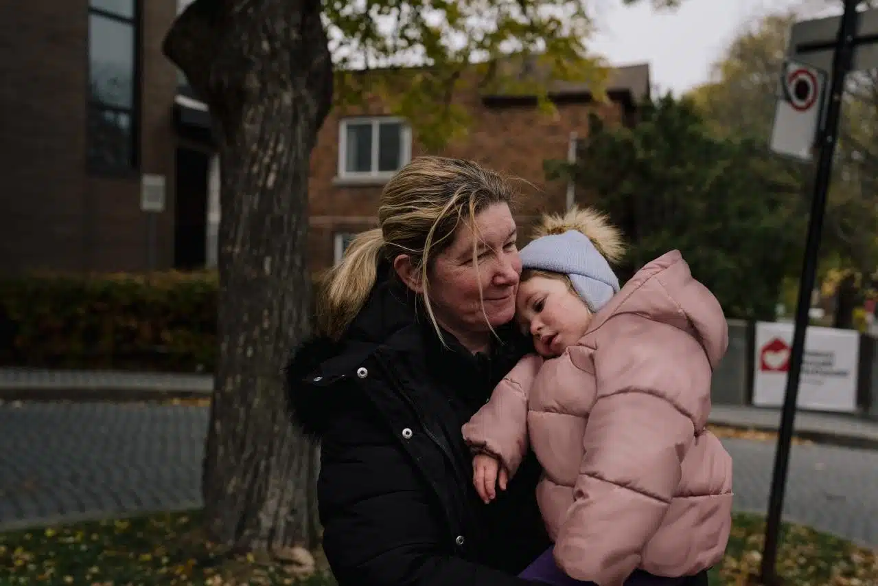 A mother in a black winter coat holds her young daughter, bundled in a pink puffer jacket and knit hat, who rests her head on her shoulder outside the brick Ronald McDonald House on an autumn day.