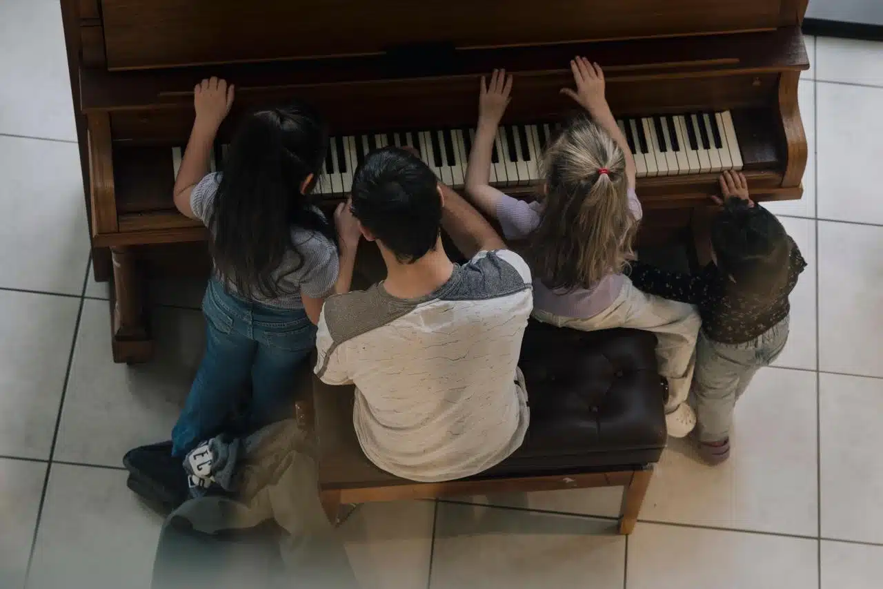 Seen from above, four children gather around an upright piano with their hands on the keys — one seated on the bench, the others standing alongside.