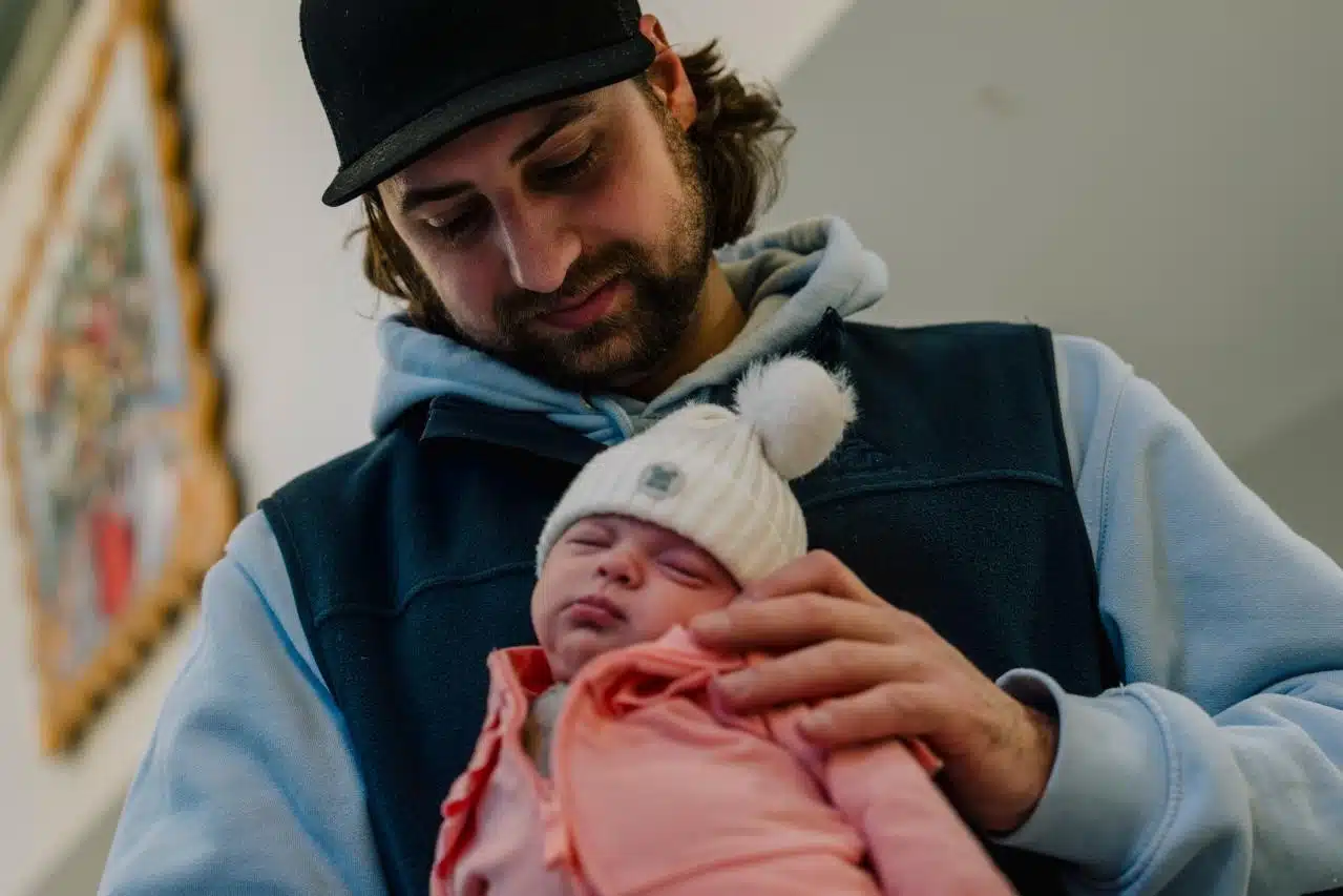 A young father in a cap gazes down tenderly at his sleeping newborn, bundled in a pink outfit and white pom-pom hat, as he cradles her against his chest.