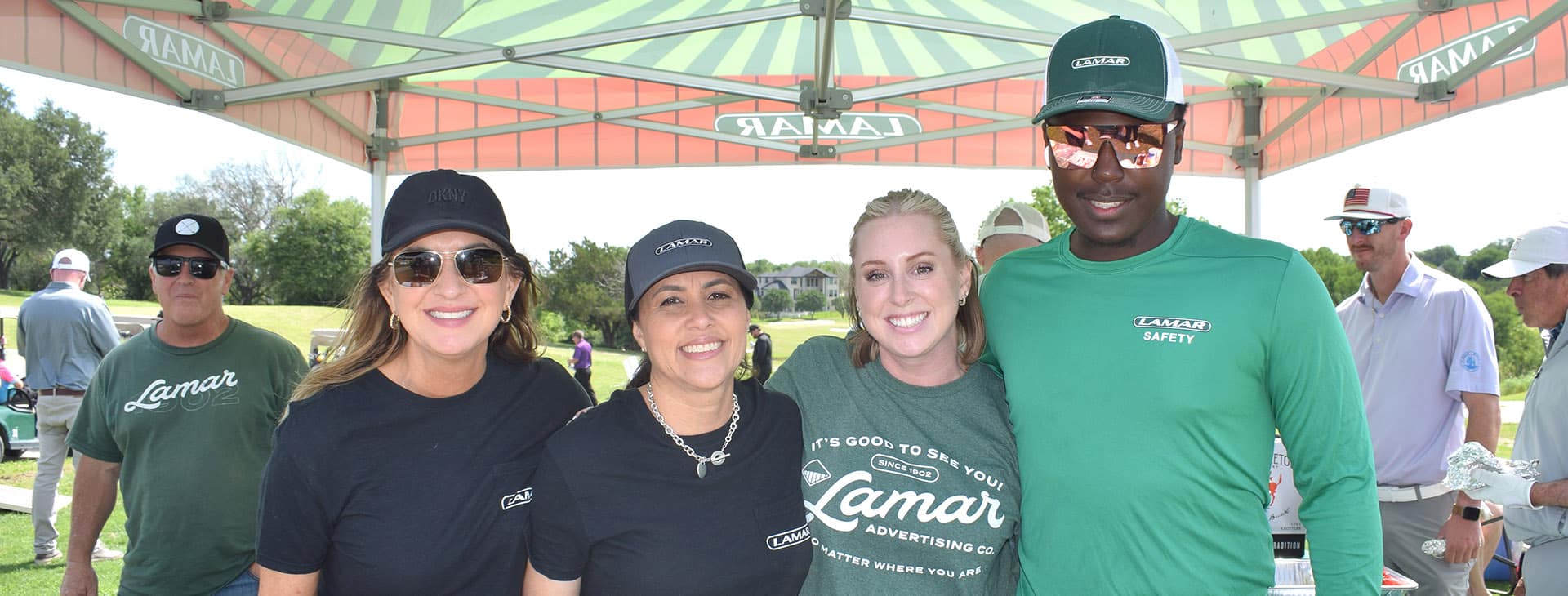 Smiling corporate volunteers in Lamar shirts and caps under a sponsor tent at a charity golf tournament
