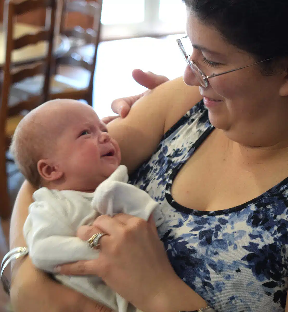 A smiling woman in glasses gently comforts a crying newborn cradled in her arms
