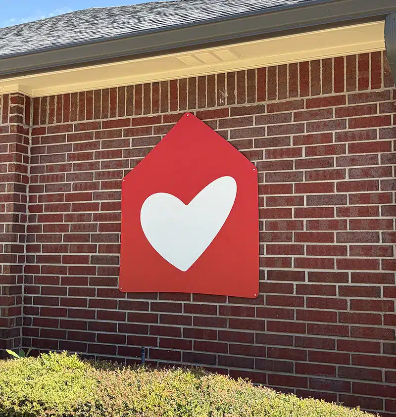 The Ronald McDonald House logo — a red house shape with a white heart in the center — mounted on the brick exterior wall of the House.