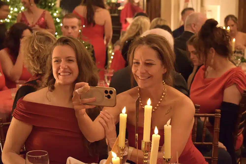 Two women in red evening dresses taking a selfie at a candlelit table during a holiday gala
