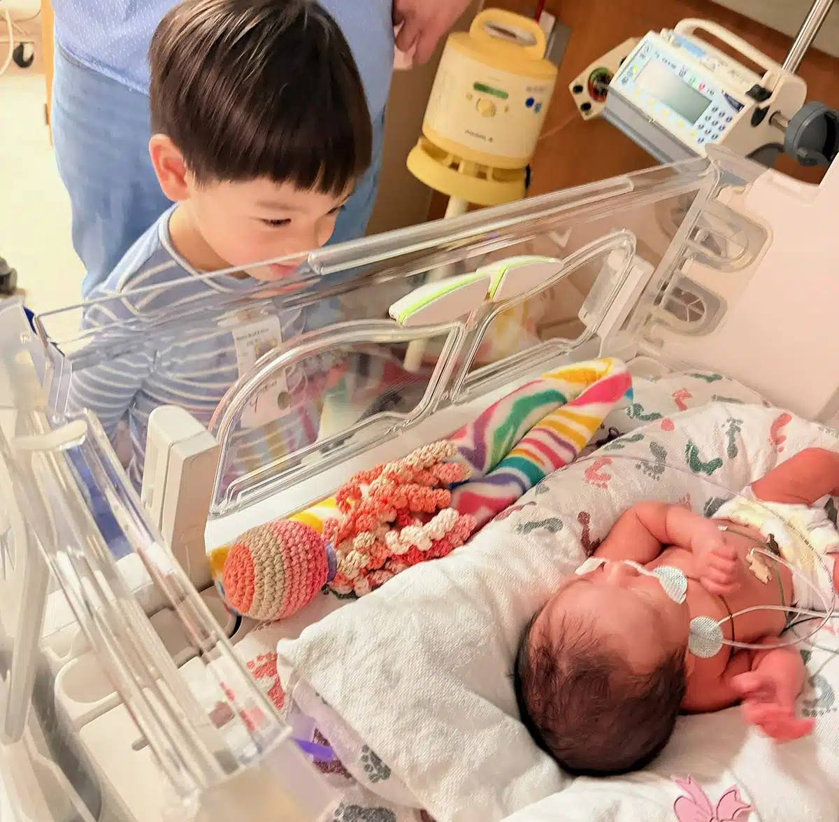 A young boy leans over a hospital bassinet to gaze at his newborn sibling in the NICU